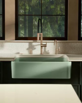 A sage green Kohler farmhouse sink with a brass kitchen faucet